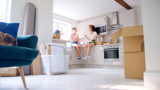 Two Female Friends Sitting On Kitchen Counter Celebrating With Wine On Moving Day In New Home