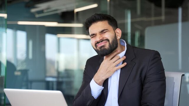 Unhealthy businessmansuffering from a sore throat and coughing sitting in business office. Sick male in formal suit has a cold, virus or flu. Unwell man holding a sore throat with his hand. Close up