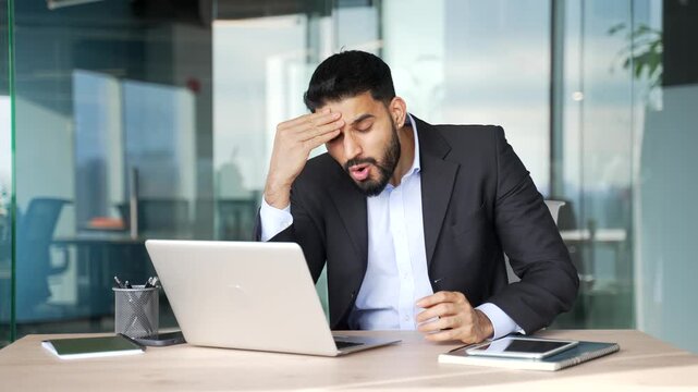 Upset frustrated businessman reads bad news on laptop while sitting at desk at workplace in modern business office. Worried male worker is sad while reviewing negative message information on computer