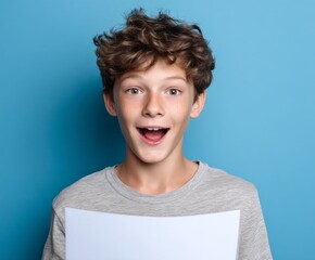 A young boy with a surprised expression holds a blank sheet of paper against a vibrant blue backdrop.