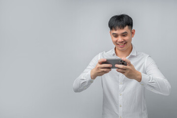 A handsome young Asian man smiles happily while using a social media or gaming app on a modern smartphone. This concept is ideal for technology, connectivity, lifestyle, and leisure.

