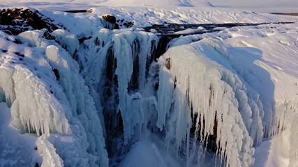 The frozen waterfall hangs with icicles as snow covers the surrounding landscape. The waterfall and icicles glisten under the sunlight on the snowy terrain. - Powered by Adobe