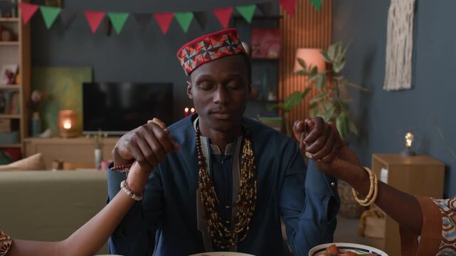 Waist up of African American man sitting with his wife and child at festive Kwanzaa table, holding hands in prayer and honoring family, tradition, and unity on this special holiday - Powered by Adobe