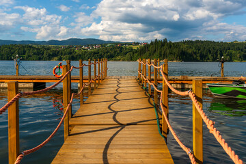 Obraz premium A wooden pier with orange rope railings casts rhythmic curved shadows on the water surface. Clear sky with fluffy clouds and forested hills create a calm, inviting summer lakeside scene.