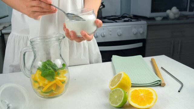 Woman preparing lemonade putting sugar to herb mint, citrus slices into glass jug. Cooking fresh homemade vitamin drink with organic products on kitchen at home. Recipe cuisine culinary ingredient.