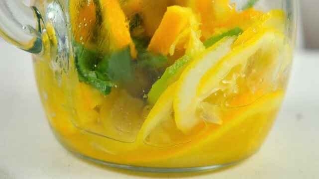 Preparing homemade lemonade mixing citrus fruit with masher stick in glass jug. Cooking mixing herb mint leaves, orange, lime, lemon with sugar on kitchen at home. Recipe cuisine culinary ingredient.