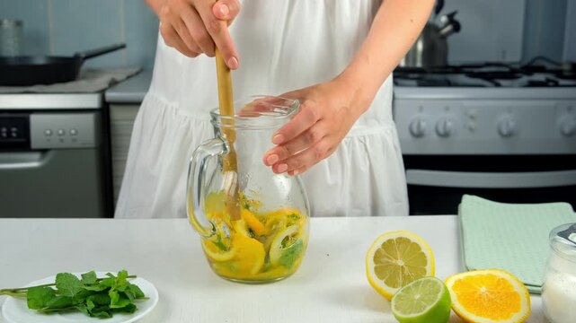Woman preparing homemade lemonade mixing citrus fruit with masher stick in jug. Cooking mixing herb mint, orange, lime, lemon with sugar on kitchen at home. Recipe cuisine culinary ingredient.