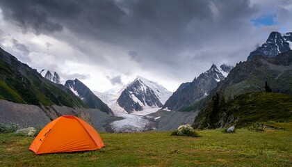 dramatic landscape with orange tent in alpine valley with view to big glacier tongue and large sharp snow capped mountain range under gray cloudy sky green hills and rocks against ice and sheer crags