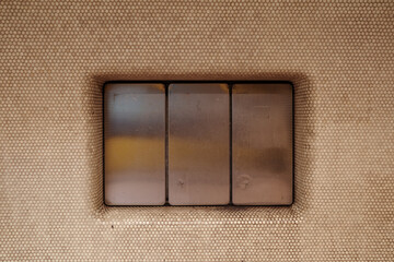 A detailed close-up of a triple-gang light switch with brushed metal buttons. The modern, minimalist switches are recessed into a beige wall panel that has a unique perforated or dimpled texture