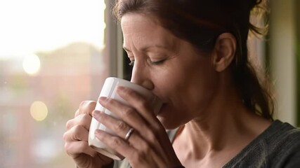 Serene middle-aged caucasian woman sips steaming white mug, gazing contemplatively at blurred city in window, warm golden hour light. Concept of peaceful morning routine - Powered by Adobe