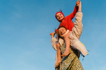 Mother carrying her daughter on her shoulder against blue sky. Smiling mom carrying happy little...