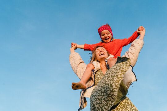 Mother carrying her daughter on her shoulder against blue sky. Smiling mom carrying happy little girl making airplane arms. Young family have fun at beach in windy winter.