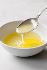 Close-up of a small white bowl on a white countertop, showing the lemon-egg mixture being stirred until smooth. The pale yellow emulsion, rich and glossy, is the key step in creating the signature cre