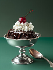 A refined studio food photography shot featuring a decadent chocolate brownie elegantly placed in a silver dessert dish. The brownie is styled with a dollop of whipped cream and topped with a glossy r