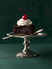 A refined studio food photography shot featuring a decadent chocolate brownie elegantly placed in a silver dessert dish. The brownie is styled with a dollop of whipped cream and topped with a glossy r