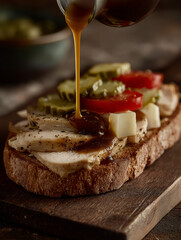 Open-face turkey sandwich on a rustic toasted bun, topped with fresh tomato slices and a thick layer of crunchy dill pickle chips. saturated Orange Sauce is being dispensed mid-motion, sauce mid-pour 