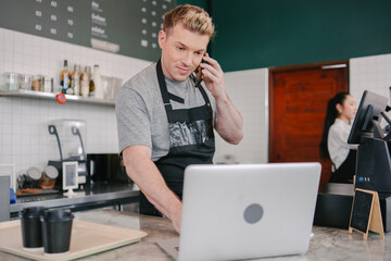 A busy male barista in an apron is talking on a smartphone and looking intently at a laptop behind...