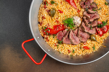 Top view of Spanish paella with juicy sliced beef steak, rice, roasted red peppers, garlic, and herbs served in a traditional pan
