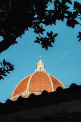The magnificent brick dome of Florence Cathedral rises majestically against a vibrant blue sky, framed by silhouetted tree leaves and a charming Italian rooftop.
