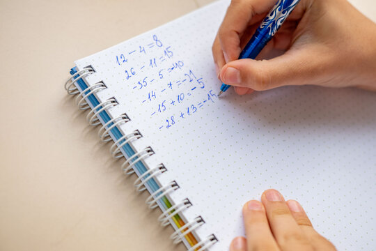 Hand writing math equations in dotted notebook with blue pen, solving arithmetic problems including negative numbers, educational activity, student homework, learning study session, school concept