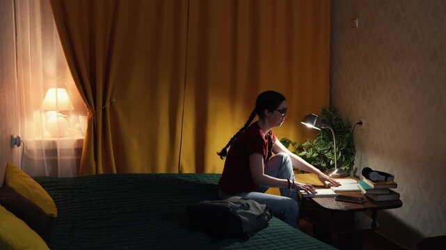 Beautiful Caucasian student woman indoors at desk studies in evening lighting working with books and laptop preparing for exams and education process. Wide shot