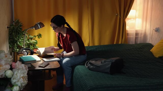 Beautiful student girl sits indoors in evening home setting at desk learning with books and laptop preparing for college exams and courses. Copy space - Powered by Adobe