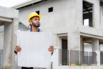 A confident construction engineer in a yellow safety helmet inspects architectural blueprints at a modern building site. He stands in front of an unfinished concrete structure under development.