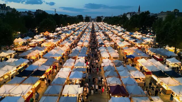 High aerial drone video of a bustling outdoor night market at dusk, featuring vibrant illuminated tents and tiny figures of people creating a dynamic urban spectacle.