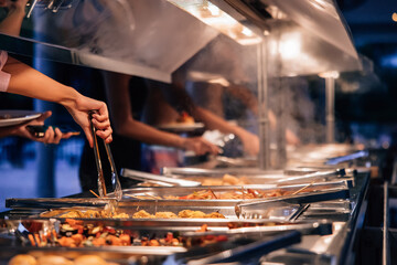 Hands of people selecting hot dishes from a steaming buffet line at an evening event, surrounded by...