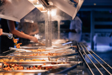 People serving skewered shrimp and various hot dishes from a steaming buffet station during an evening catered event or dinner