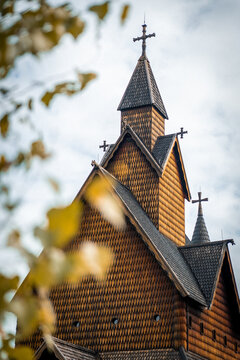 Close-up on the unique, historic architecture of a typical Norwegian Stave Church, captured under a cloudy sky. The roof is covered with traditional brown-orange wooden shingles, topped with crosses a