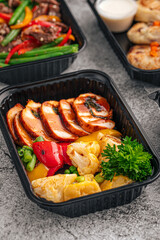 Sliced grilled chicken breast with colorful vegetables and herbs in a black lunchbox on gray background