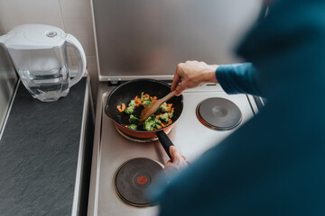 A person prepares vegetables in a frying pan on a stove top in their kitchen, emphasizing healthy cooking and home cooking. A water filter jug is visible on the kitchen countertop nearby.