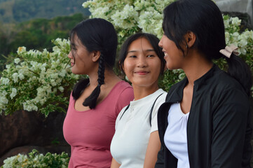 Three teenage girls sit close together by the temple garden edge, dressed in traditional skirts and laughing happily with blooming white flowers creating a bright background.