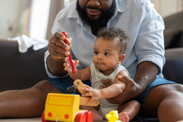 African American dad helps his baby play with colorful wooden toys while sitting together on the floor in home.