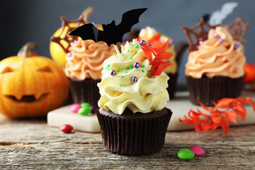 Delicious cupcake with bat topper, sprinkles, branch with red leaves and colorful candies on wooden table against grey background, closeup. Halloween celebration