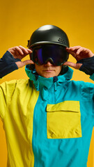 Skier adjusting goggles in yellow studio portrait with ski jacket. Concept of winter sportswear campaigns, protective eyewear focus, professional athlete imagery and fashion-oriented advertising.