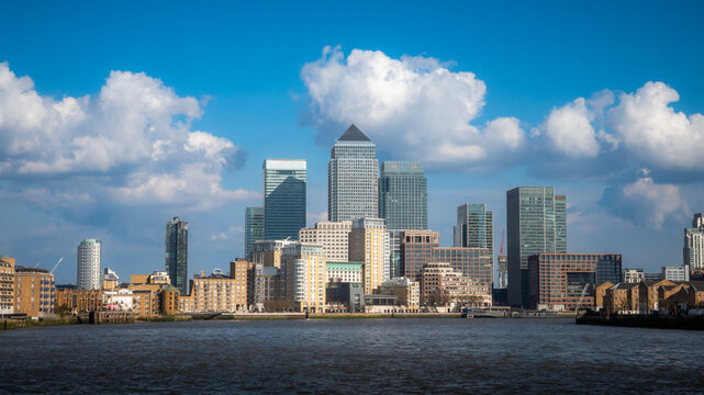 Docklands, London. The imposing skyscrapers of Canary Wharf dominate the skyline across the River Thames on a clear day with scattered white clouds and bright sunshine.