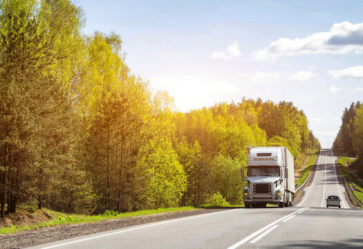 American semi-trailer truck transports cargo along country road uphill against sun and forest in summer. Copy space for text, logistics company