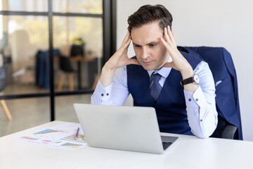 Stressed businessman in formal attire sitting at desk, concentrating on laptop screen with financial charts, illustrating workplace pressure and data analysis stress.
