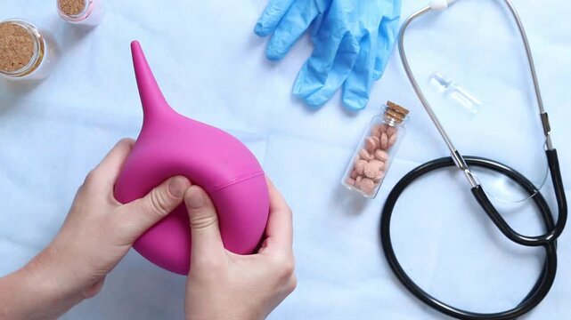 Doctor's hands holding a large pink enema on a table background with various objects, top view. Female doctor squeezing a pink rubber enema. Nurse or doctor's desk with a stethoscope, pills and gloves