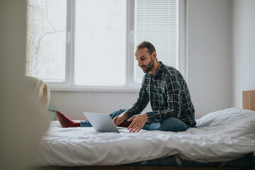 A young man in casual attire sitting cross-legged on his bed, focused on working with a laptop....