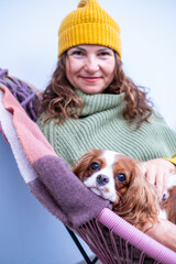 Midlife woman in cozy green sweater and yellow hat relaxes on balcony with dog, enjoying autumn warmth and companionship while wrapped in a blanket, embodying comfort and serenity