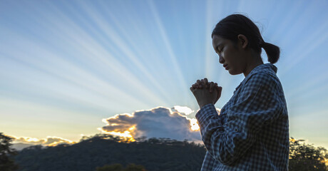 A girl praying to God with holding hands and closing her eyes at sunset shining brightly sky background.
