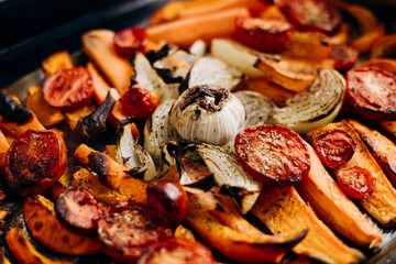 Roasted autumn vegetables pumpkin tomato onion garlic on baking tray, close up.