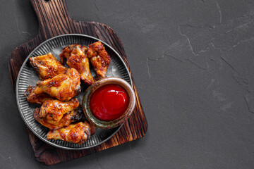 Grilled spicy chicken wings with ketchup in a plate on a dark slate, stone or concrete background. Top view with copy space.