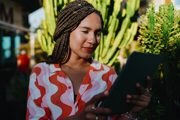 Young woman with braids holding digital tablet outdoors, focused and confident, symbolizing empowerment, freelance independence, and creativity through modern technology. © BullRun
