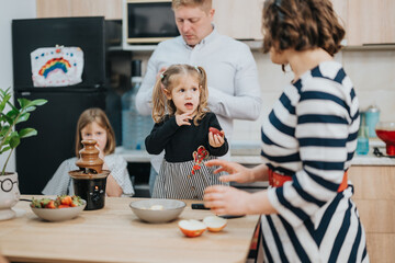A heartwarming moment of a family gathering in the kitchen preparing and enjoying a chocolate fondue with fruits, showcasing togetherness, bonding, and joyful interaction in a warm home environment.