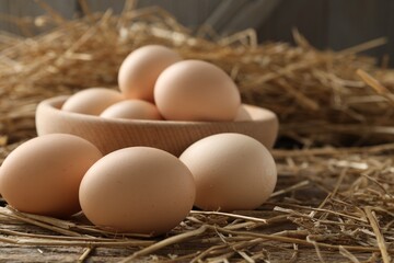 Raw chicken eggs in bowl and straw on wooden table, closeup