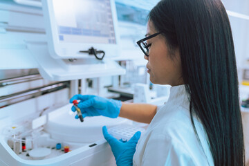 Female lab technician in blue protective gloves putting test tubes with blood samples in a biochemistry centrifuge analyzer.